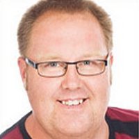 Der Mann hat eine fröhliche Ausstrahlung, trägt eine Brille und hat einen leichten Bart, während er in die Kamera lächelt.