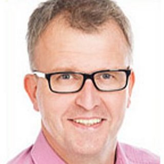 Ein lächelnder Mann mit Brille und kurzem, grauem Haar trägt ein rosa Hemd und strahlt Freundlichkeit aus.