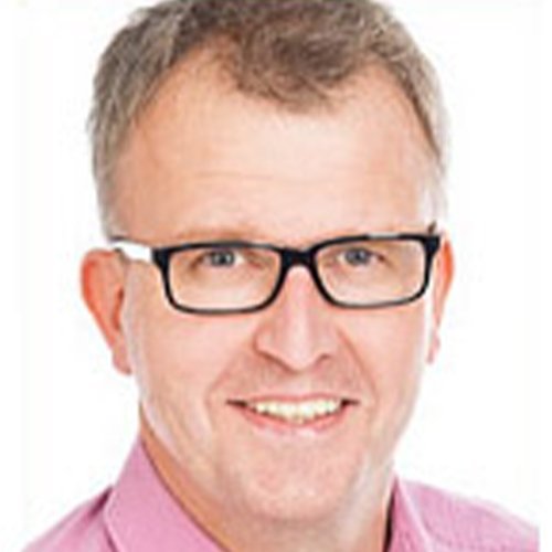 Ein lächelnder Mann mit Brille und kurzem, grauem Haar trägt ein rosa Hemd und strahlt Freundlichkeit aus.