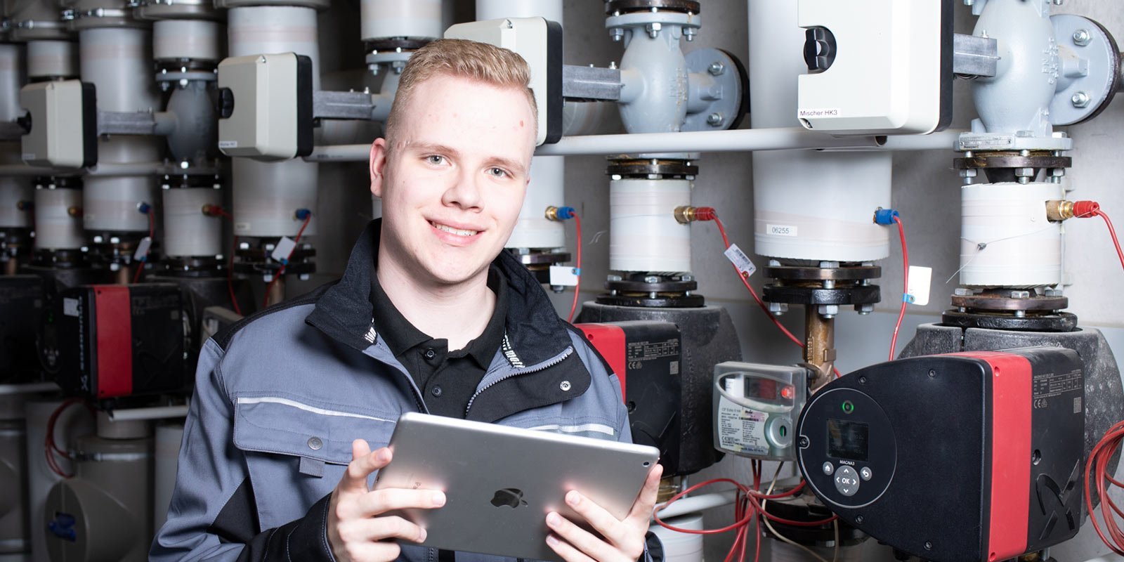 Ein junger Anlagenmechaniker vor einer Heizungsanlage, der ein Tablet hält 
